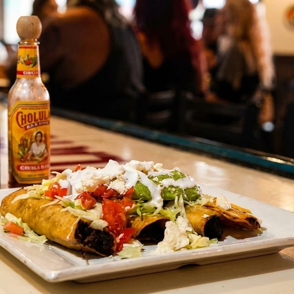 Three crispy flautas topped with guacamole, sour cream, and salsa, with hot sauce bottles on a bar.